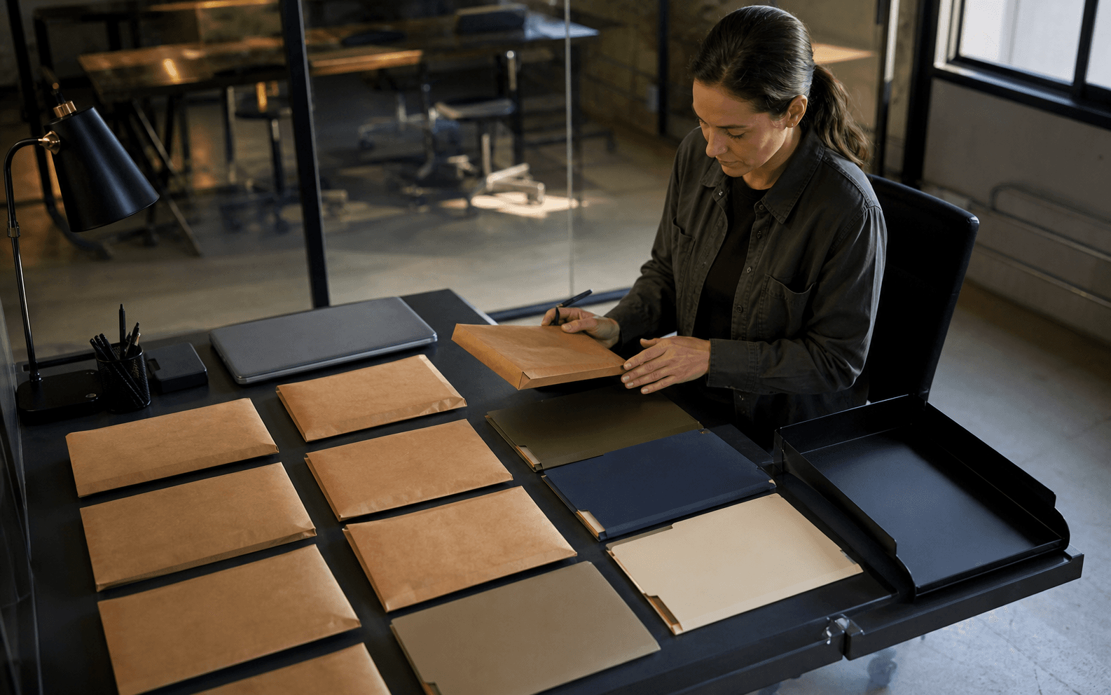 Production administrator reviewing separated sealed packets and blank color-tabbed folders in a glass-partitioned office